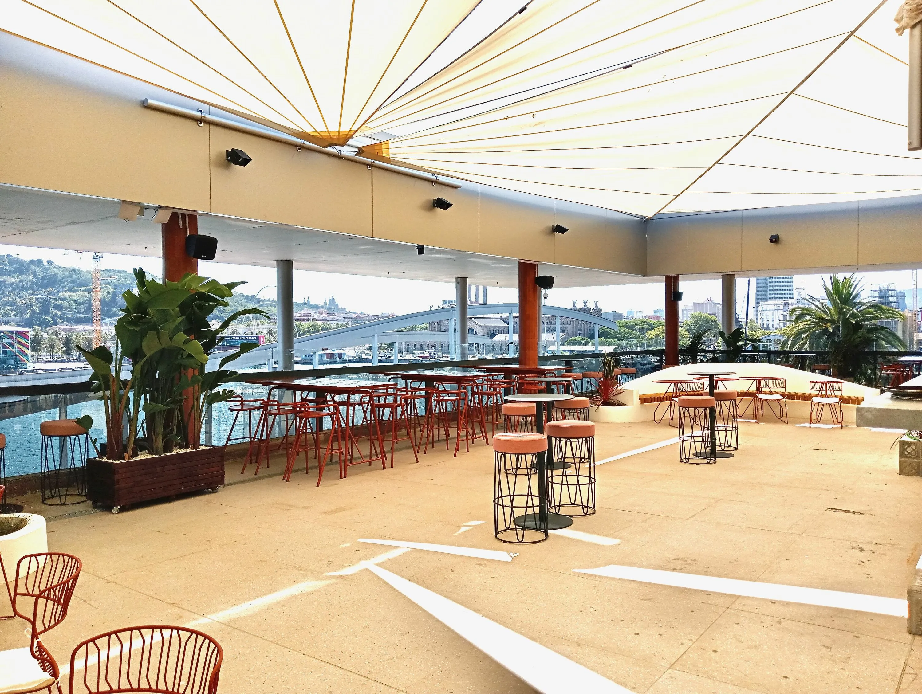 Bar central con diseño abierto y sofisticado, rodeado de mesas modernas bajo velas tensadas y vistas al puerto. Ideal para copas con brisa marina.