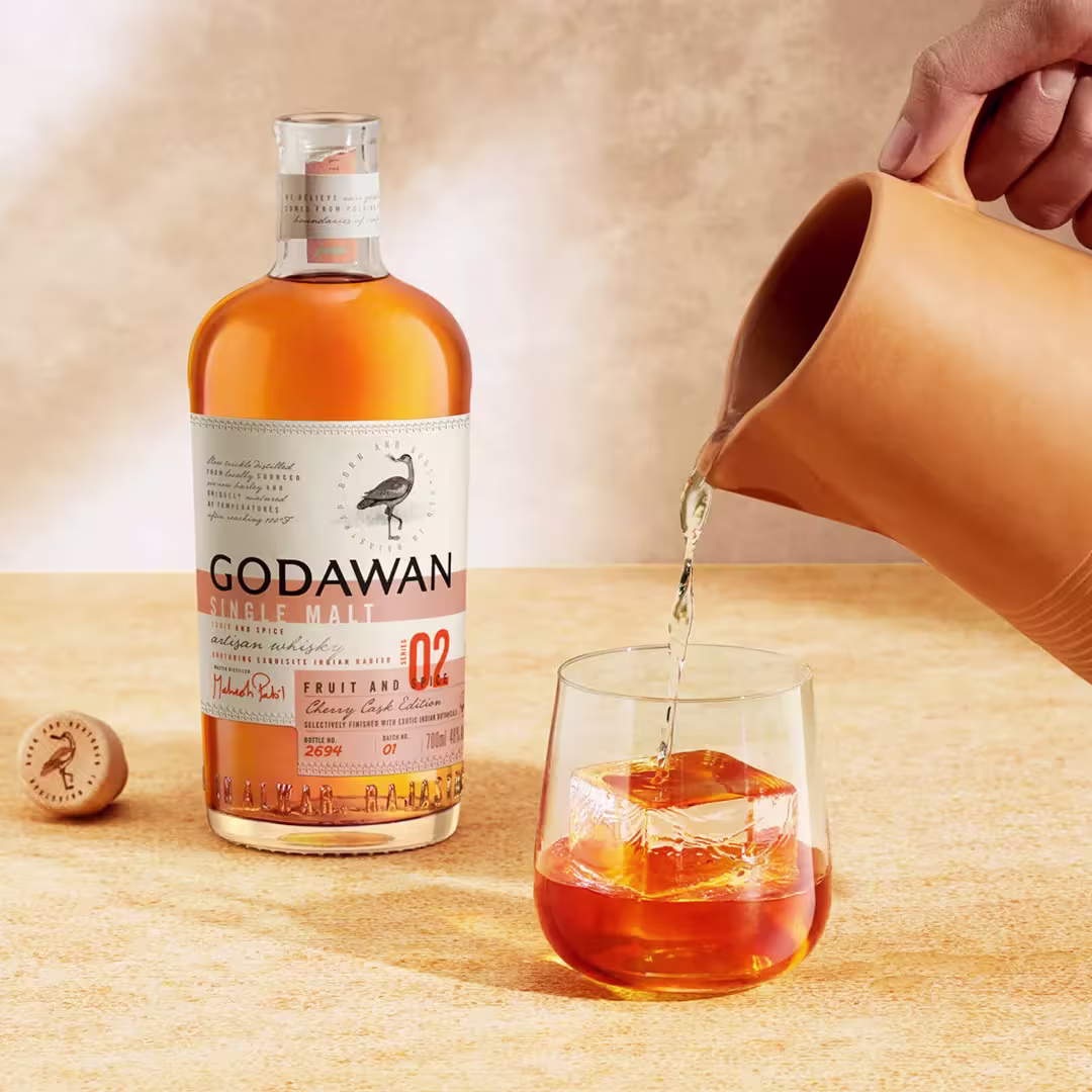 A hand pours liquid from a ceramic pitcher into a glass with ice, next to a bottle of Godawan Single Malt whisky on a beige surface.