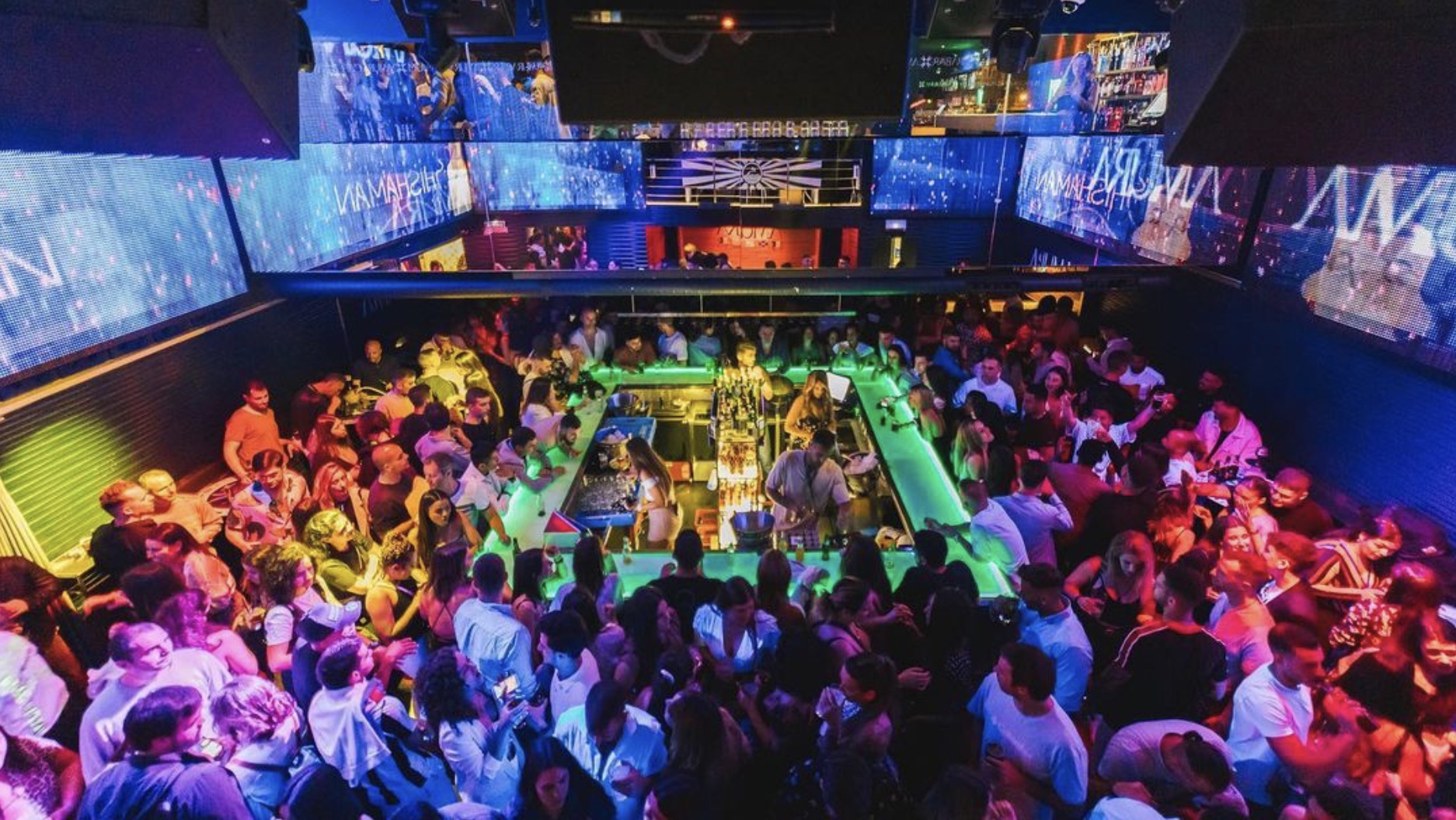 Interior de Amura, con una barra central iluminada en verde, pantallas LED, y un ambiente lleno de energía con una multitud disfrutando la música.
