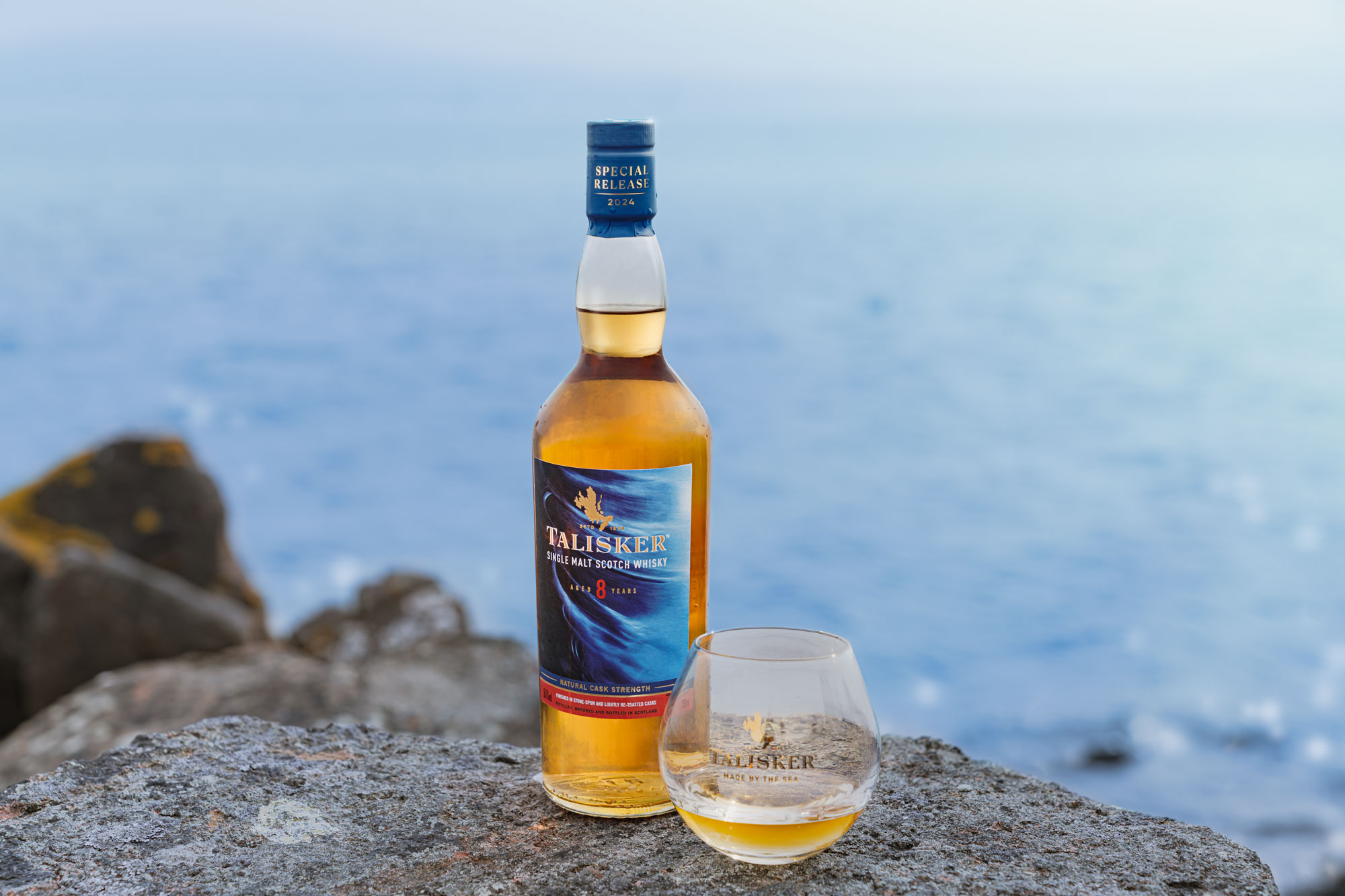A bottle of Talisker whisky and a glass with whisky sit on a rocky surface by the sea, with blue water and rocks in the background.