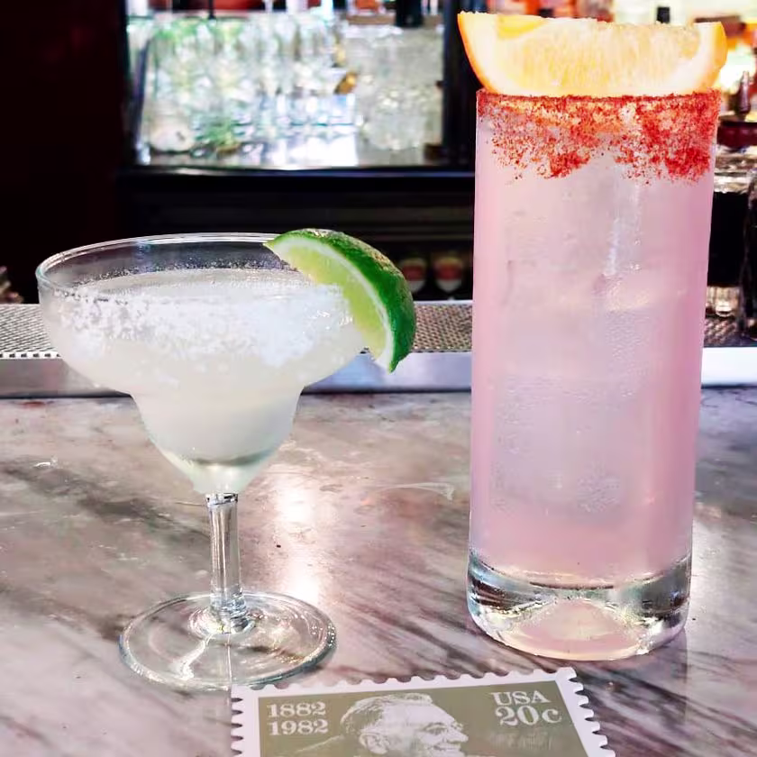 Dos tragos en primer plano, distintos vasos. Una estampa de época como posa vasos. Barra del bar de fondo.