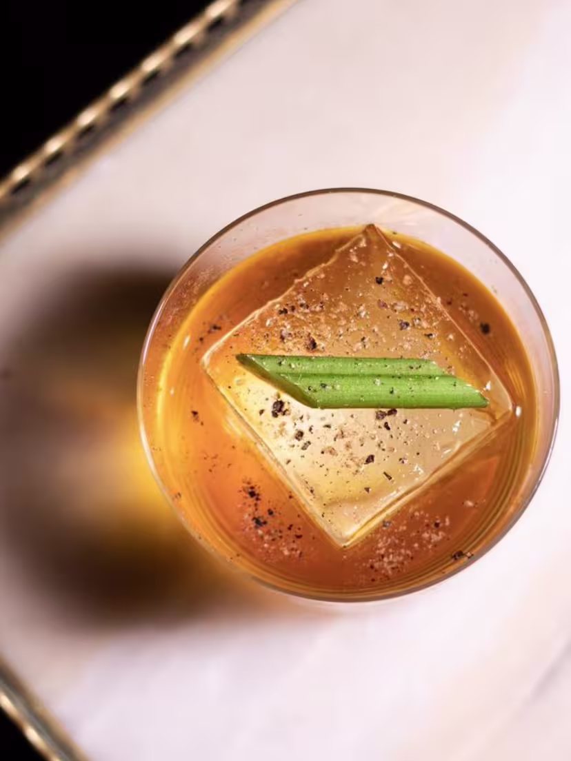 En Santamaría Coctelería, un cóctel dorado se presenta con un cubo de hielo perfecto, decorado con una hoja verde y un toque de especias.