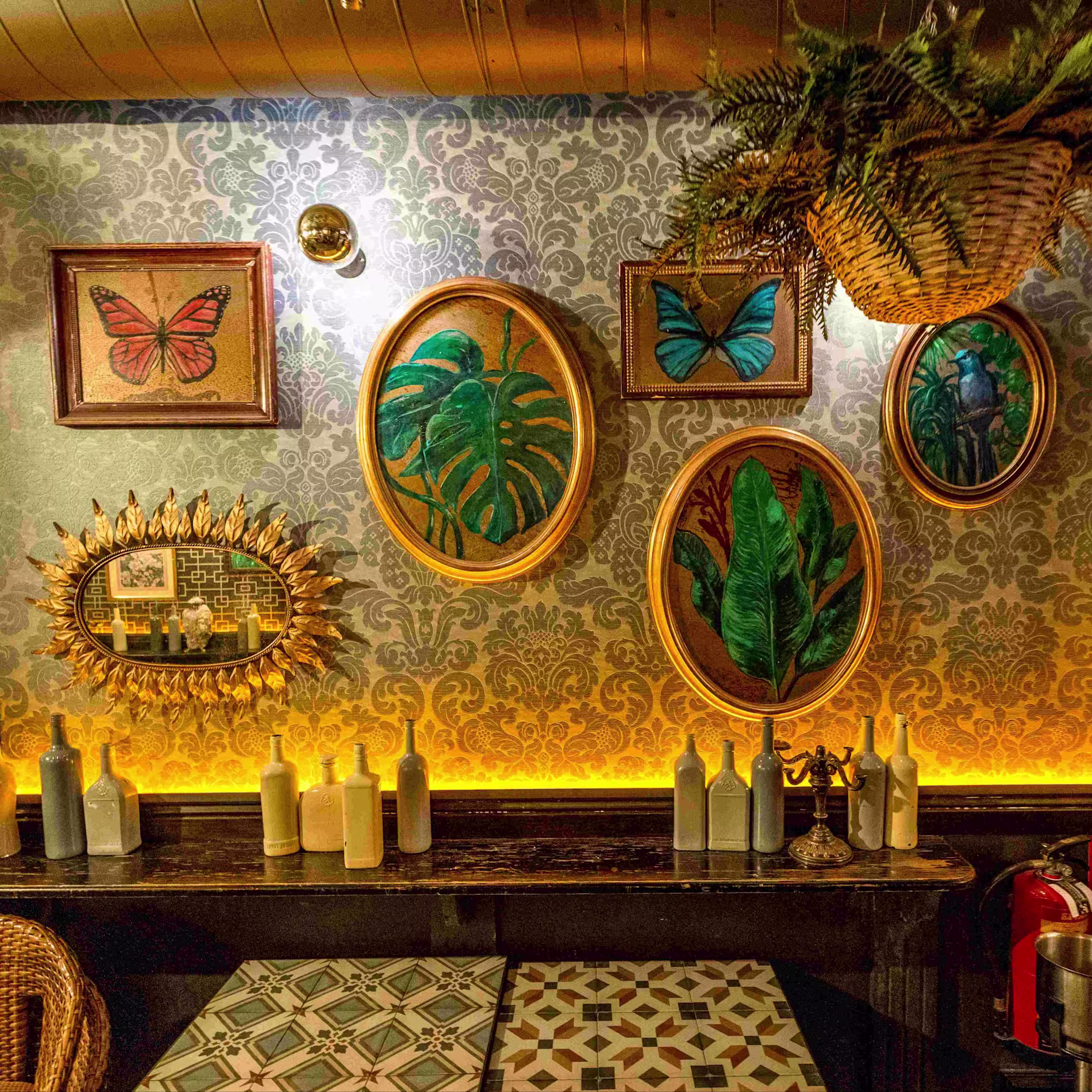 Pared empapelada con estampado floral, cuadros de hojas y mariposas, botellas decorativas y luz cálida que resalta cada detalle. Un rincón acogedor de Lola 09.