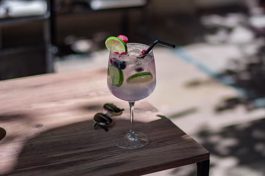 A glass of clear cocktail with ice, lime slices, berries, and a black straw sits on a wooden table in an outdoor setting.