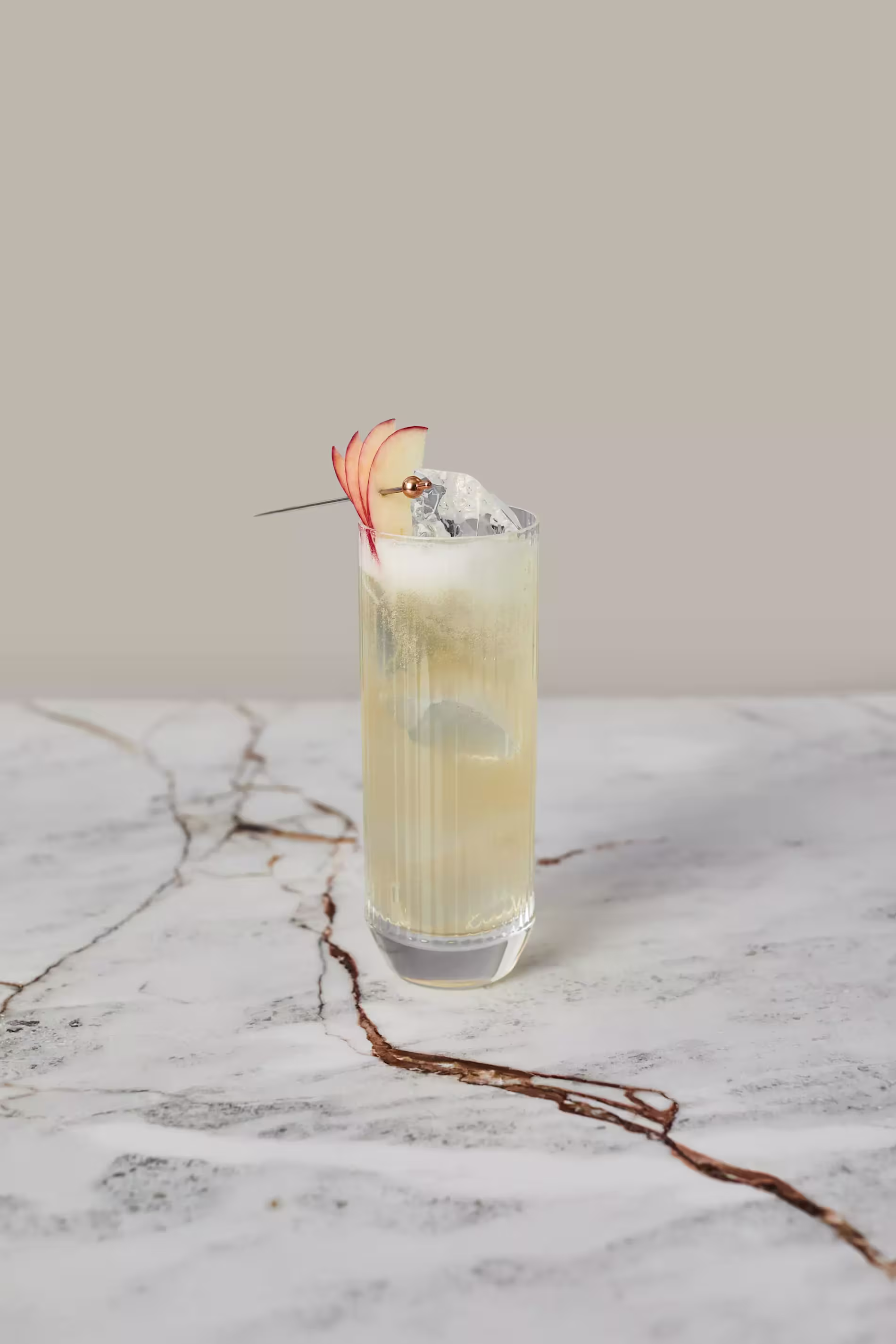 A tall glass of light-colored cocktail with ice cubes, garnished with thin apple slices and a cherry on a skewer, sits on a marble surface.