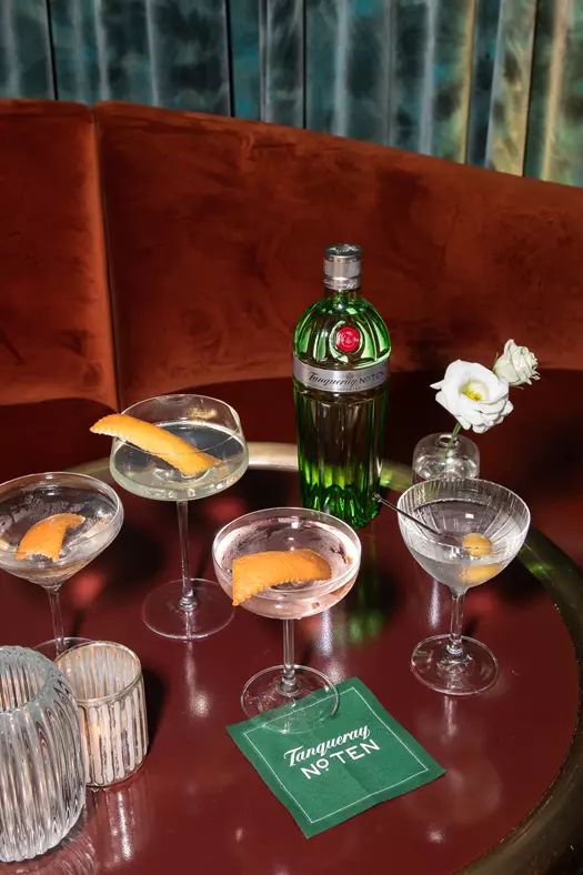 A bottle of Tanqueray No. Ten gin is on a round table with four cocktails garnished with citrus peels, a white flower in a vase, candles, and a branded napkin in a lounge setting.