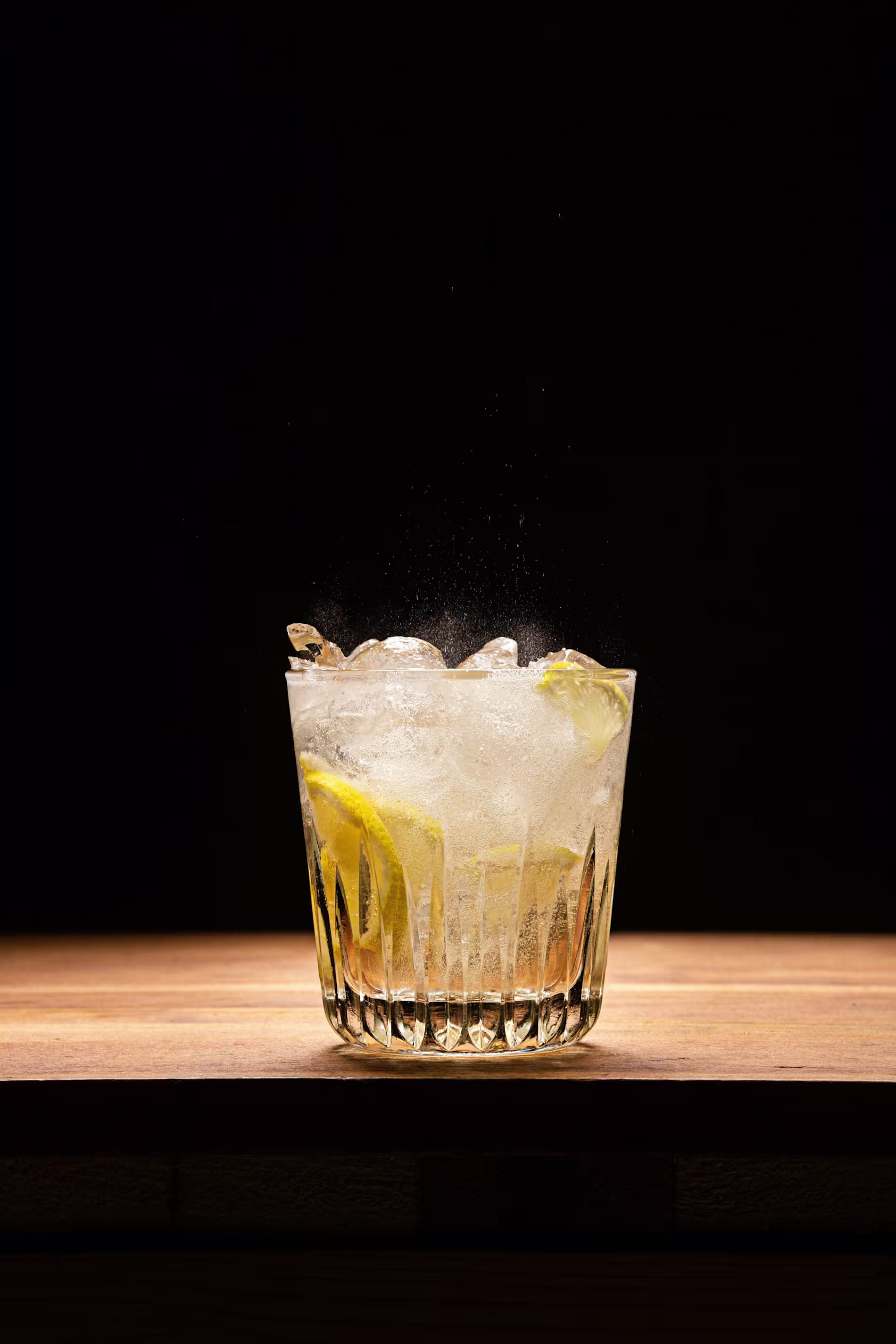 A glass of clear iced drink with lemon slices sits on a wooden surface against a dark background.