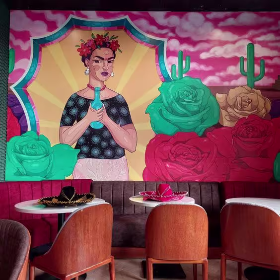 En la imagen se ve un mural que decora el bar Rosa Mezcal, y las mesas a su alrededor. Decora en la mesa unos gorros mexicanos .