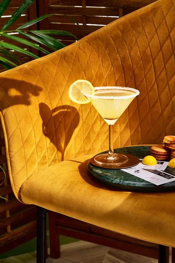 A cocktail glass with a sugared rim and lemon slice sits on a green tray atop a mustard-colored velvet chair, next to several macarons and some small papers.