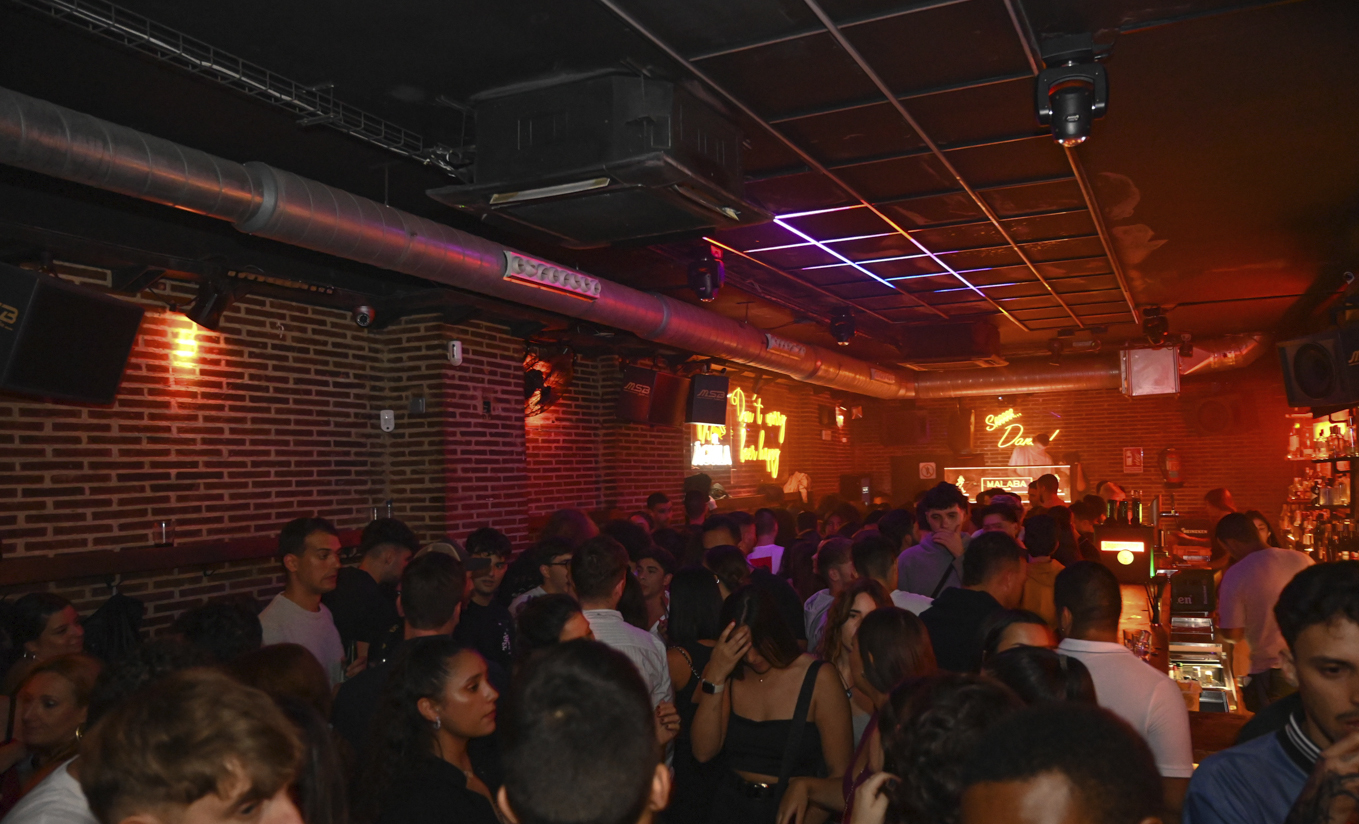 La imagen muestra el interior de Malaba Club, un espacio lleno de gente disfrutando de la noche. Con paredes de ladrillo, iluminación tenue y luces de neón que crean un ambiente moderno e industrial.