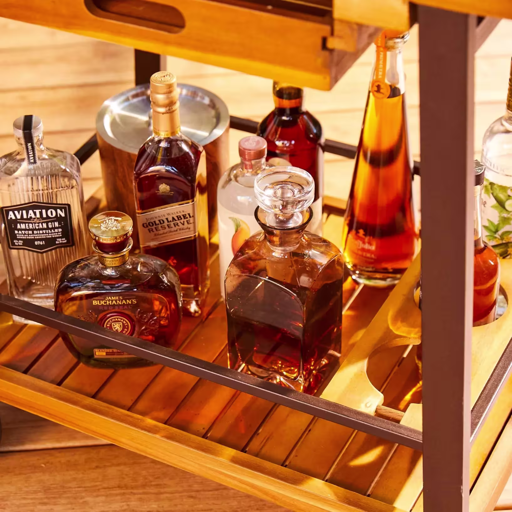 A wooden bar cart shelf holds assorted bottles of liquor, including whiskey, gin, and vodka, along with a glass decanter and wooden wine holder.