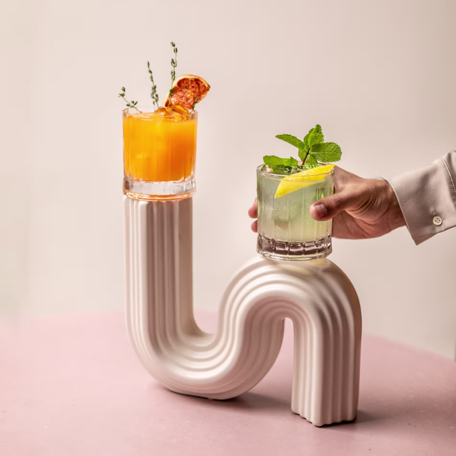 La foto muestra una configuración creativa y visualmente interesante donde se colocan dos bebidas en una estructura curva, blanca y en forma de S. A la izquierda, hay un cóctel naranja y una rodaja de fruta en el borde.
