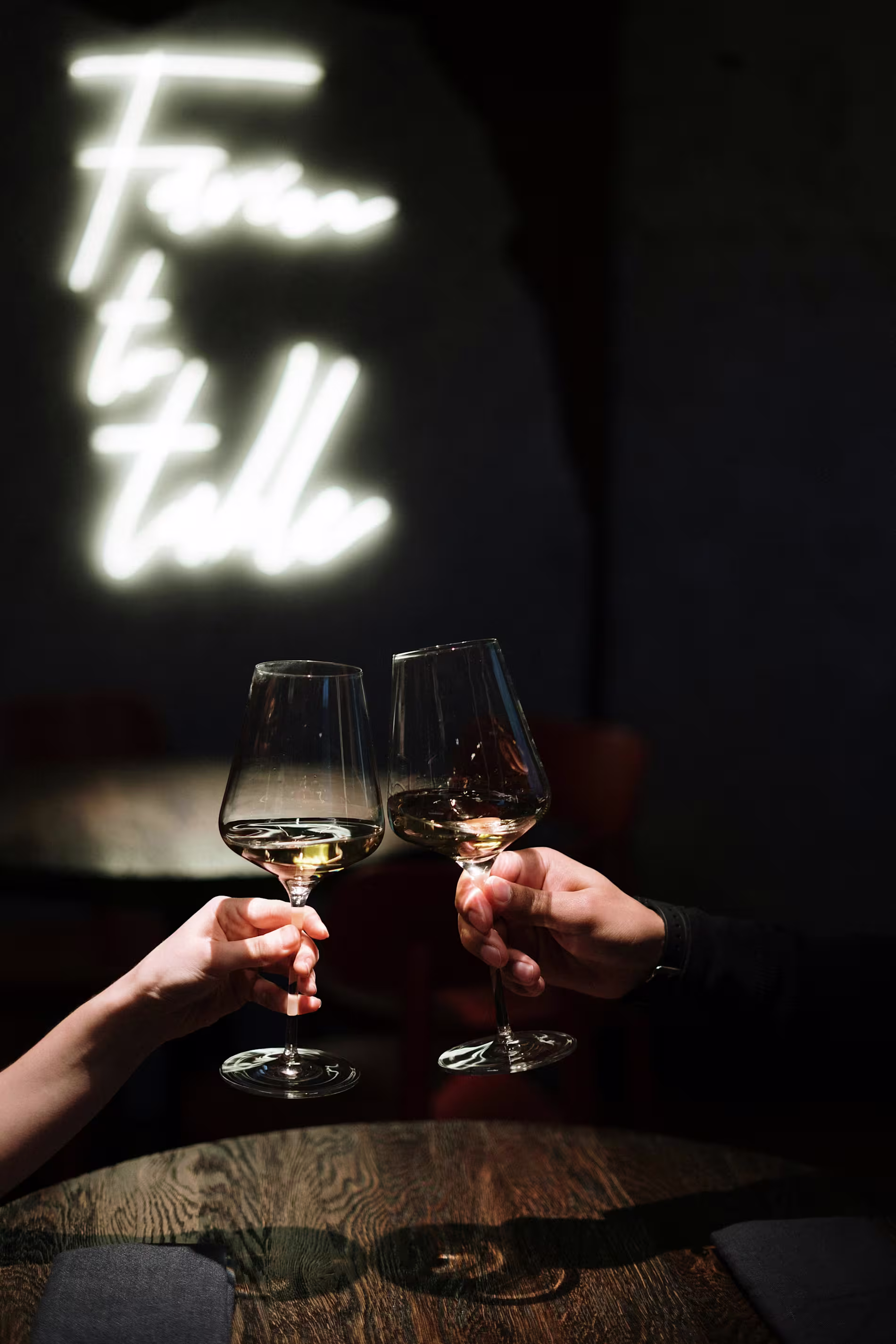Two people clink wine glasses over a dark wooden table, with a neon Farm to table sign glowing in the background.