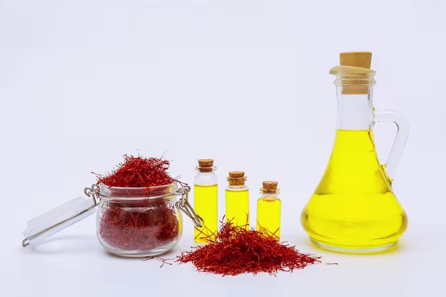 A glass jar and piles of saffron threads sit beside bottles and a carafe of yellow oil, all on a white background.