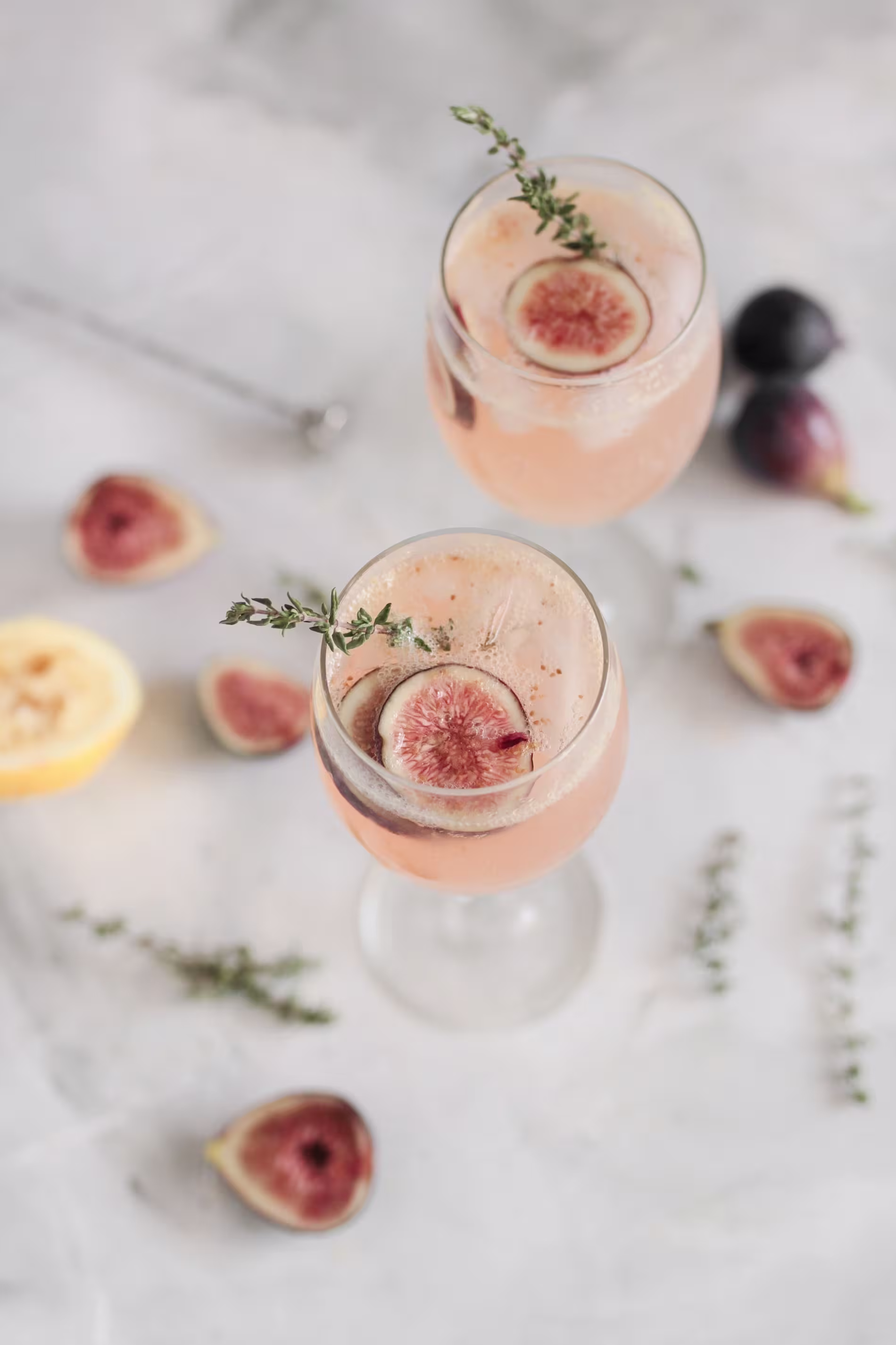 Two glasses of a pale pink cocktail garnished with fig slices and thyme sprigs, surrounded by cut figs, a lemon half, and thyme on a light surface.