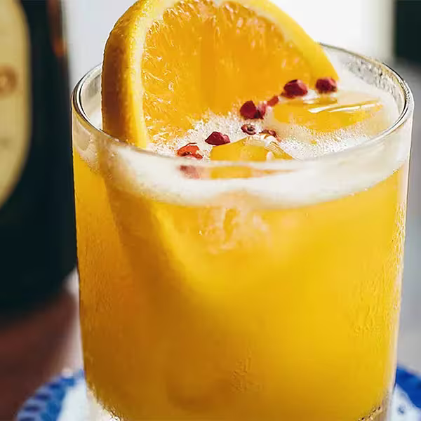 A glass of orange-colored drink with ice cubes, garnished with an orange slice and red peppercorns.