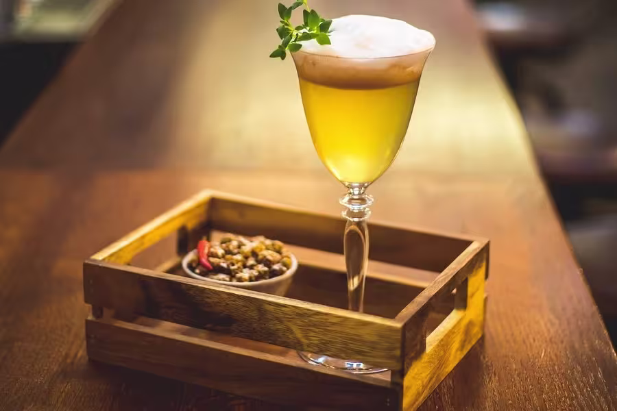 A yellow cocktail with foam, garnished with a green herb, sits in a stemmed glass next to a small bowl of snacks in a wooden tray on a wooden surface.