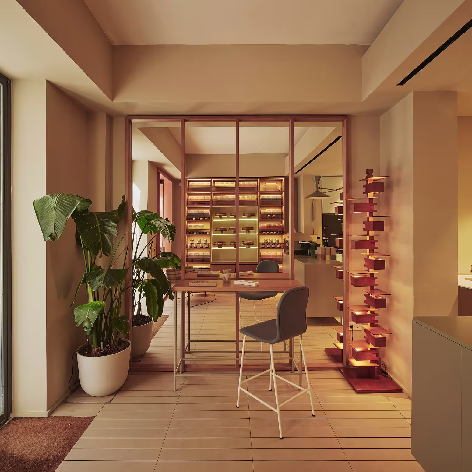 Una mesa con sillas altas junto a una planta de interior y vitrinas de madera iluminadas llenas de productos en Luna & Wanda.