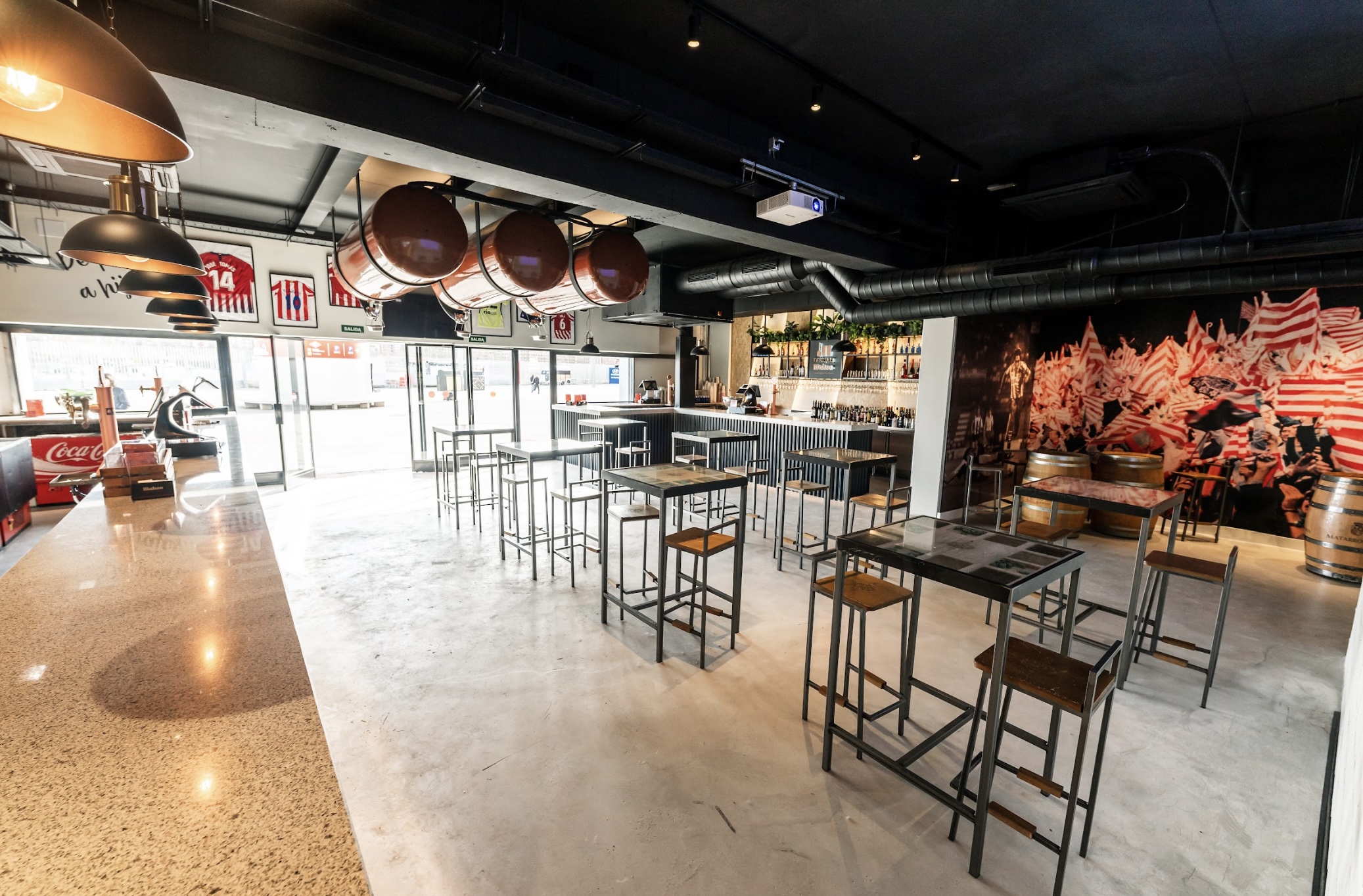 La imagen muestra el Brindis Bar, ubicado en el estadio del Atlético de Madrid. Con un diseño moderno, presenta mesas altas, decoración con camisetas del equipo y un mural con hinchas en celebración. 