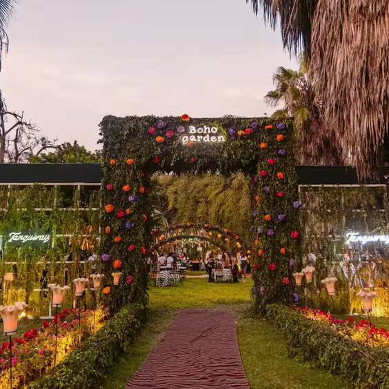 En la imagen se ve la entrada al evento, un marco verde decorado con flores, el logo de Boho garden en blanco, y carteles LED de ambos lados con logos Tanqueray
