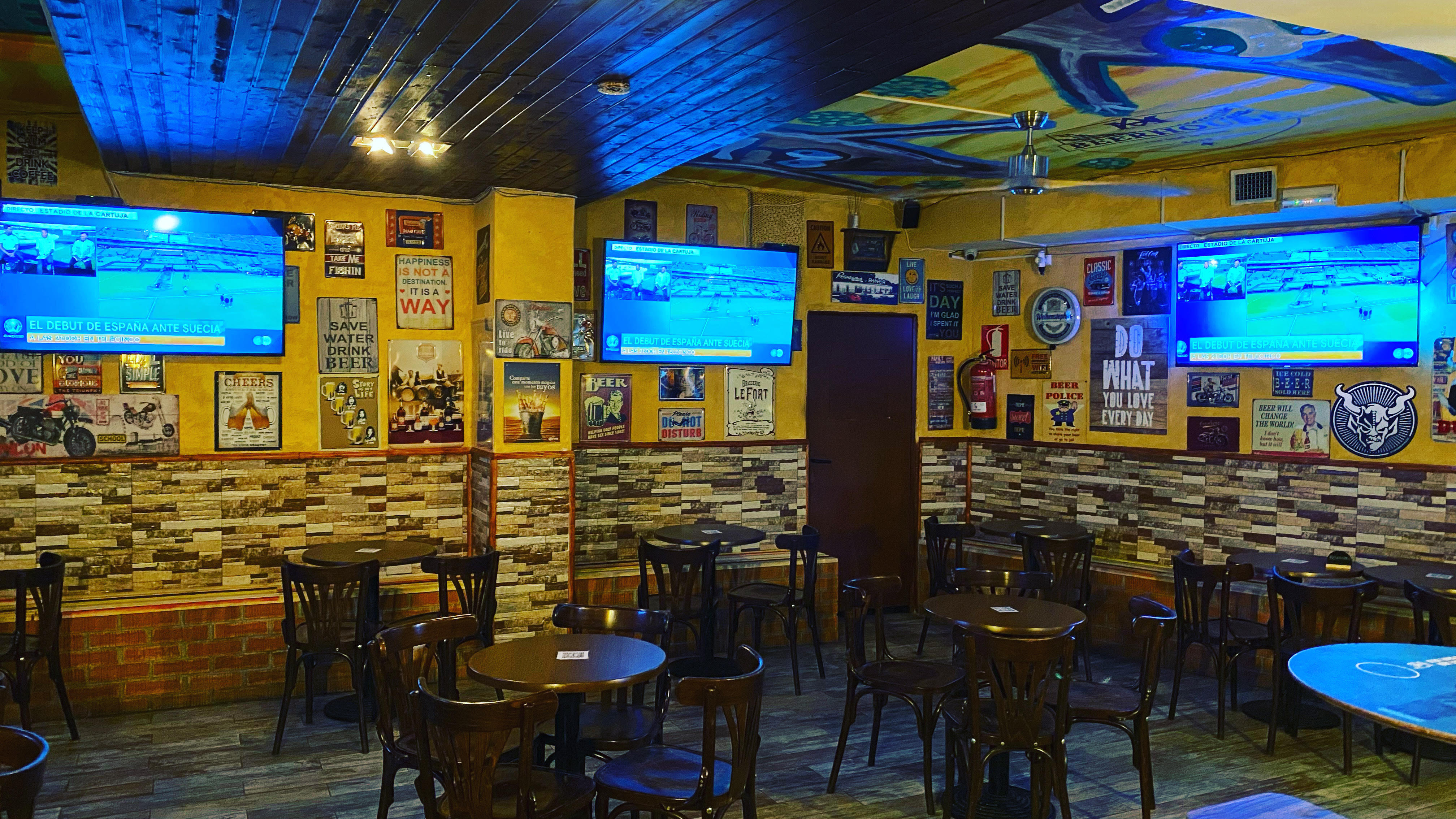 La imagen muestra el interior de Beerhouse. Hay varias mesas y sillas dispuestas para los clientes, con las paredes decoradas con diversos carteles y pósteres. El techo está pintado en un tono azul con algunos diseños, y hay varias televisiones.