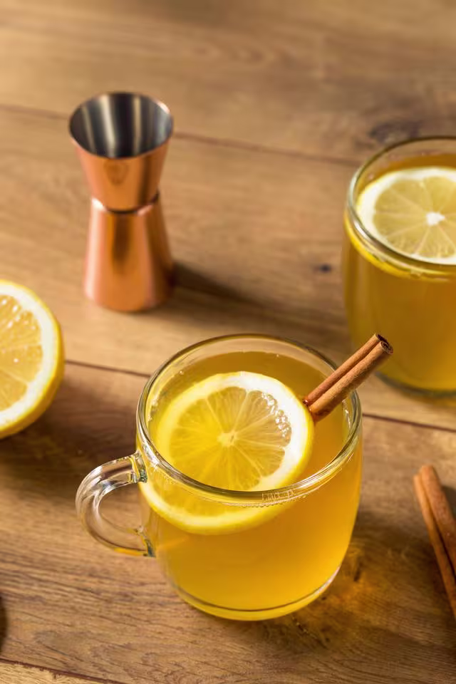 A glass mug of hot toddy with a lemon slice and cinnamon stick on a wooden table, with a jigger and another mug in the background.