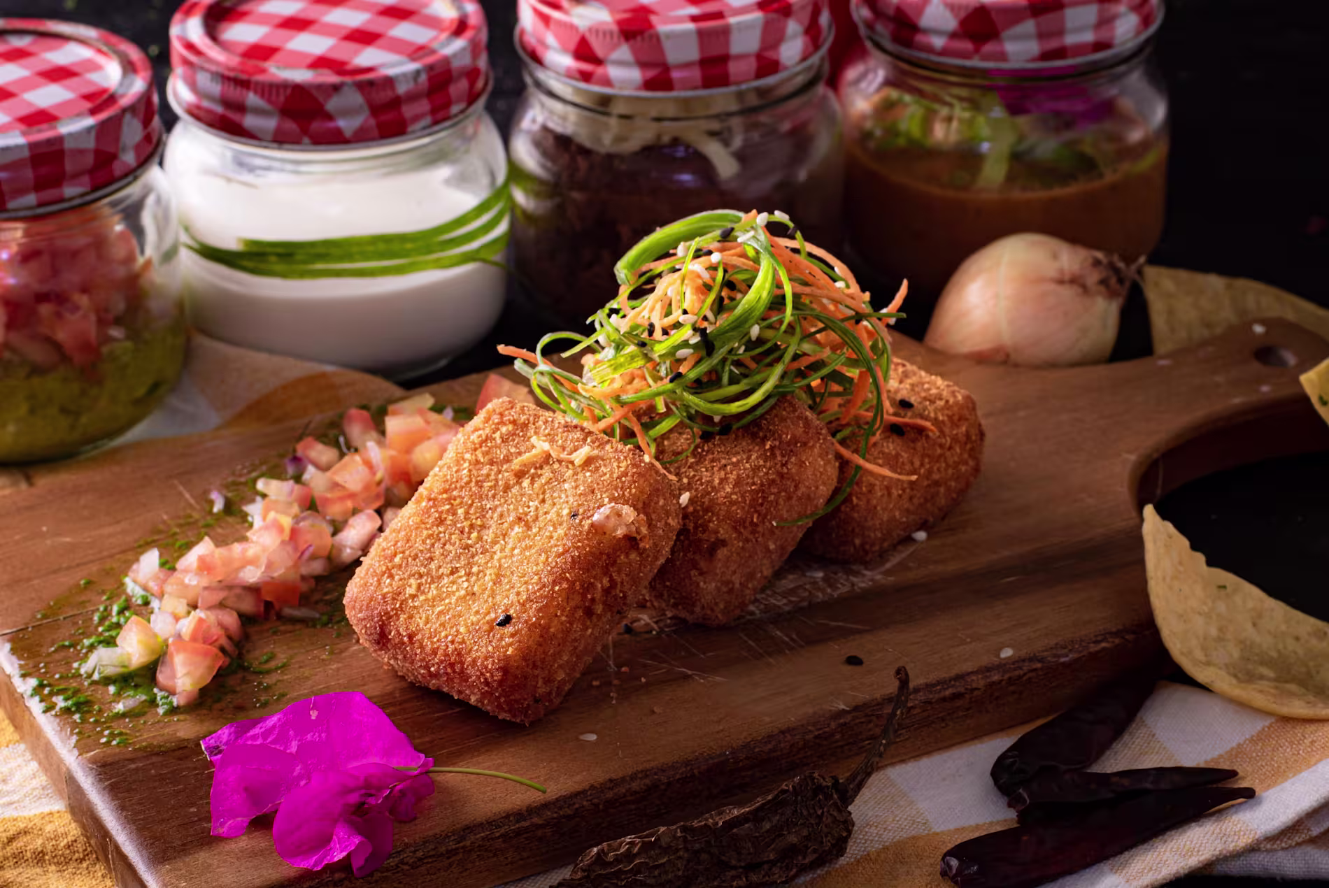 Three breaded, fried cubes topped with shredded vegetables sit on a wooden board beside a small pile of diced salsa, with jars of condiments and an onion in the background.