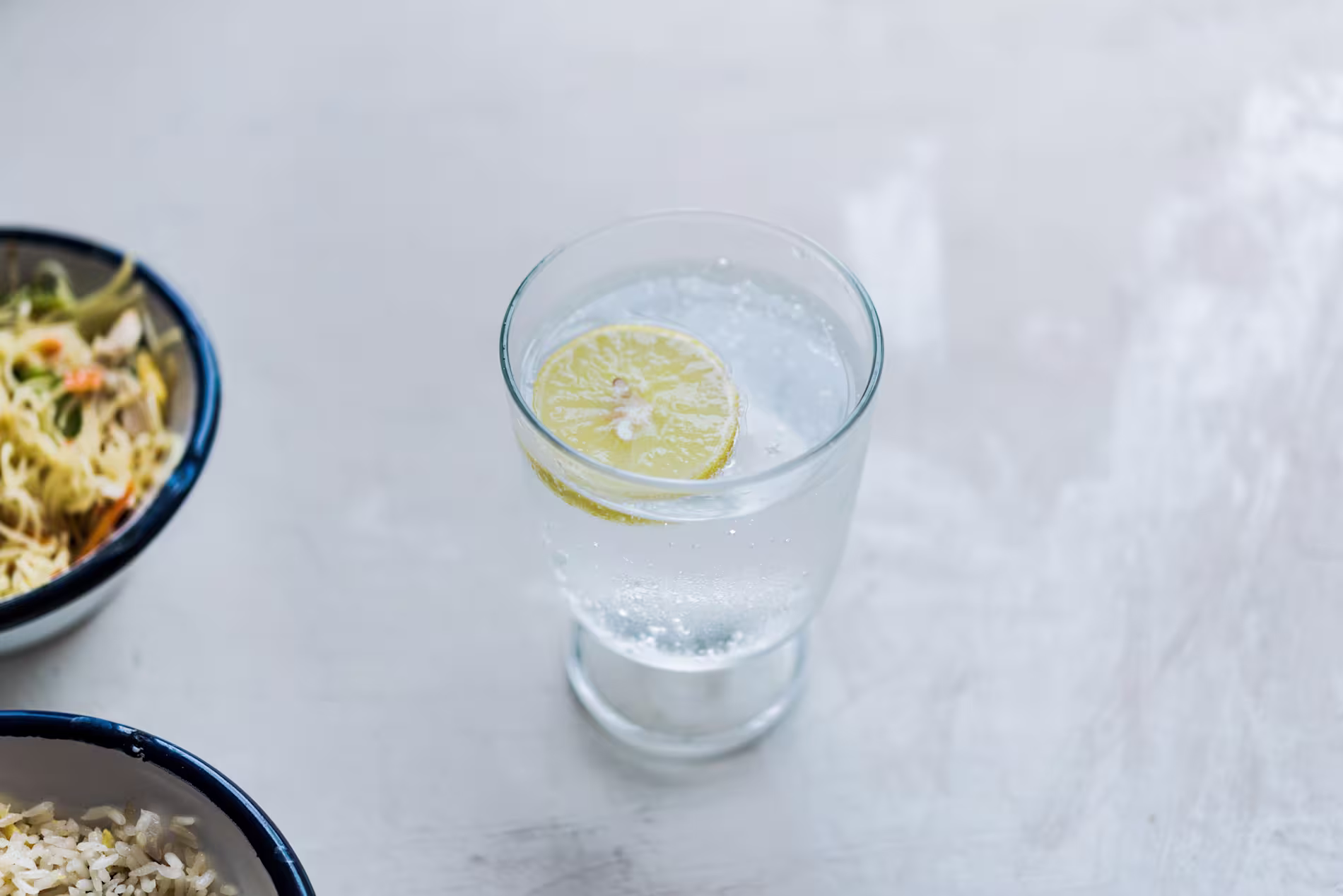 A glass of sparkling water with a lemon slice on a light surface, with bowls of food partially visible nearby.