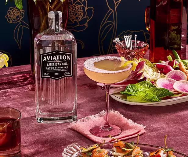 A bottle of Aviation American Gin stands next to a cocktail in a coupe glass, surrounded by plates of salad and appetizers on a pink tablecloth.