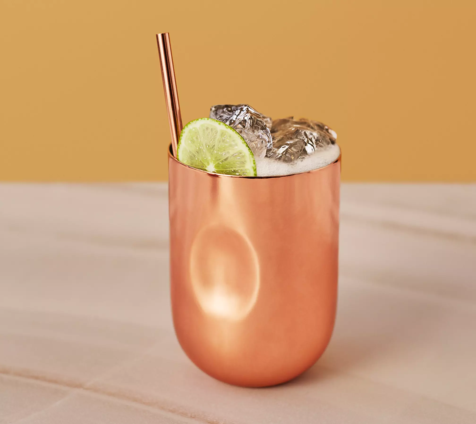 A copper cup filled with a foamy drink, ice cubes, a lime slice garnish, and a copper straw, set against a beige and yellow background.