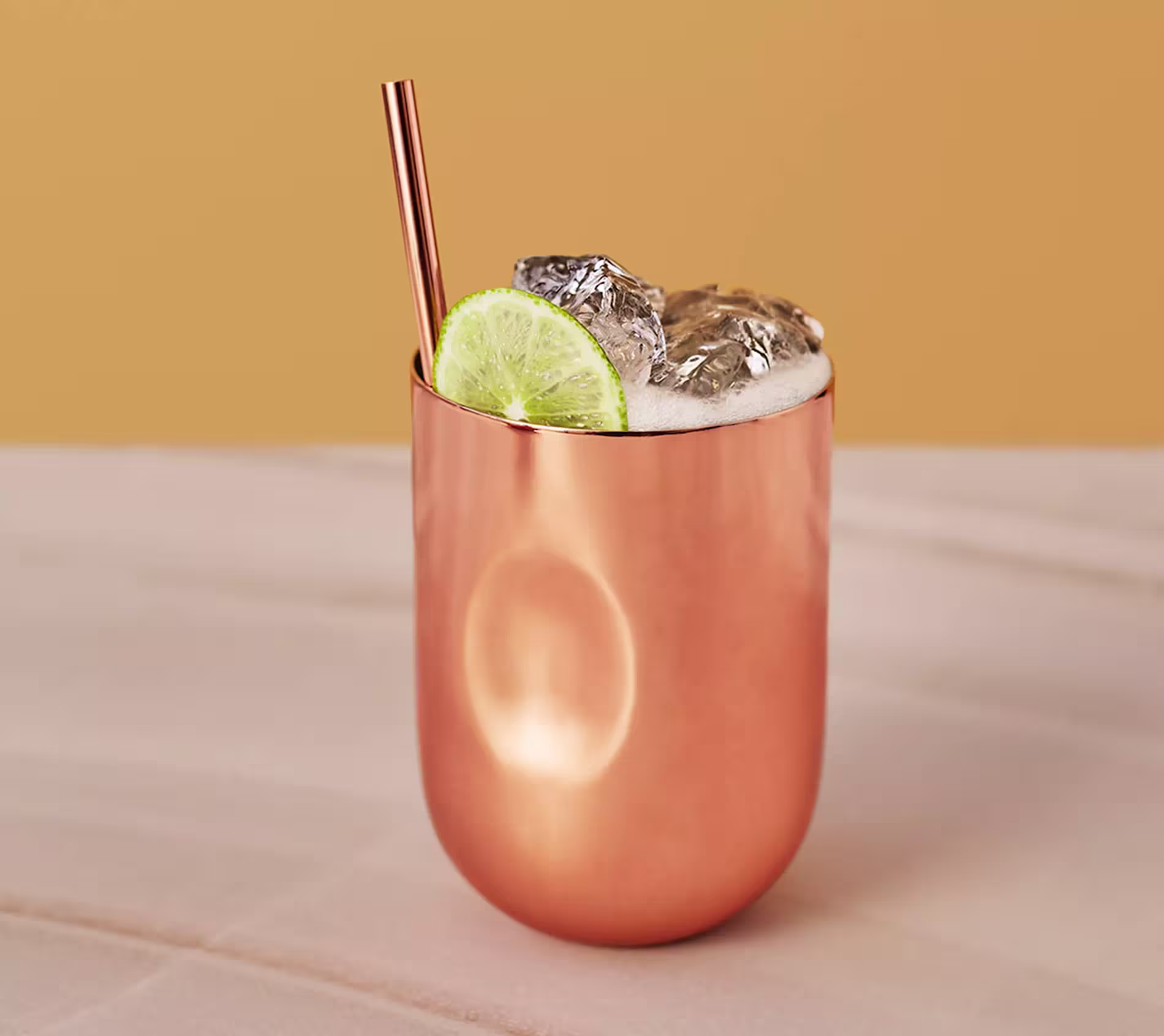 A copper cup filled with a foamy drink, ice cubes, a lime slice garnish, and a copper straw, set against a beige and yellow background.