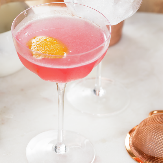 La imagen muestra una copa rellena con el cóctel Cosmopolitan, una rodaja de limón y otros elementos de coctelería.