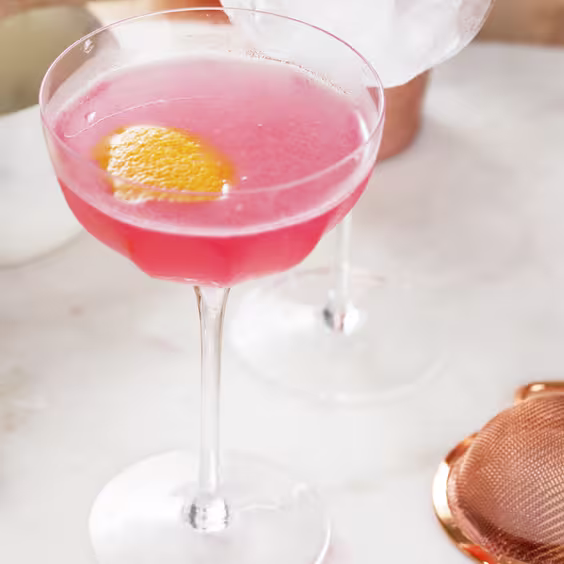 La imagen muestra una copa rellena con el cóctel Cosmopolitan, una rodaja de limón y otros elementos de coctelería.