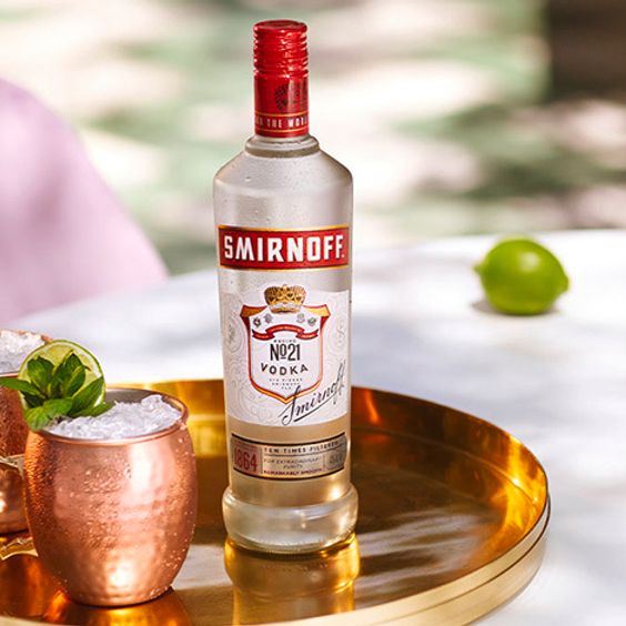 La imagen muestra una botella de vodka Smirnoff N°21 junto a dos vasos de cobre con el cóctel Moscow Mule sobre una bandeja dorada.