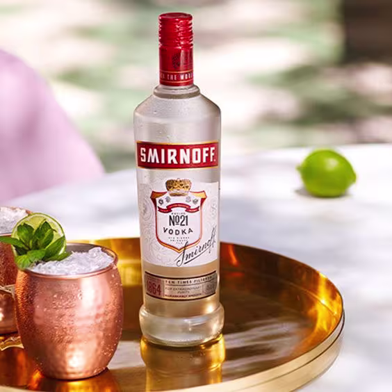 La imagen muestra una botella de vodka Smirnoff N°21 junto a dos vasos de cobre con el cóctel Moscow Mule sobre una bandeja dorada.