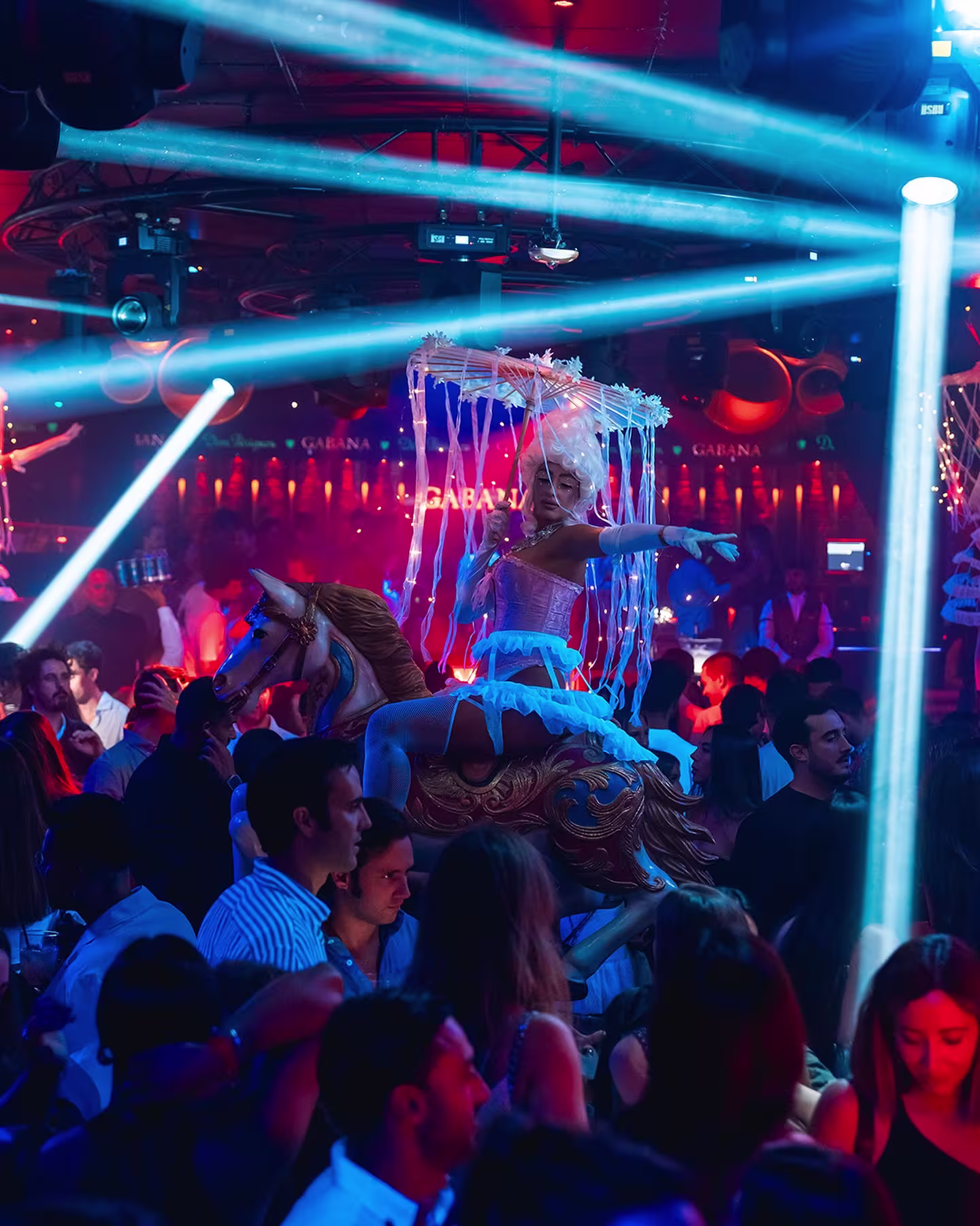 Gabana Club: Un espacio vibrante y lleno de energía, con una bailarina en traje llamativo sobre un caballo de carrusel, rodeada de luces intensas y una multitud animada.