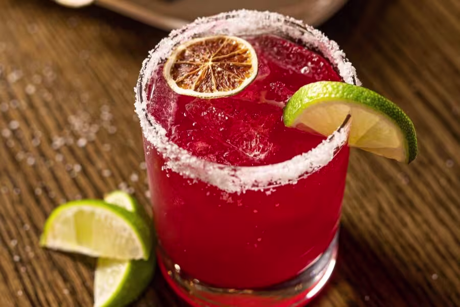 A red cocktail in a glass with a salted rim, garnished with a dried lime slice and a fresh lime wedge, on a wooden table with lime wedges beside it.