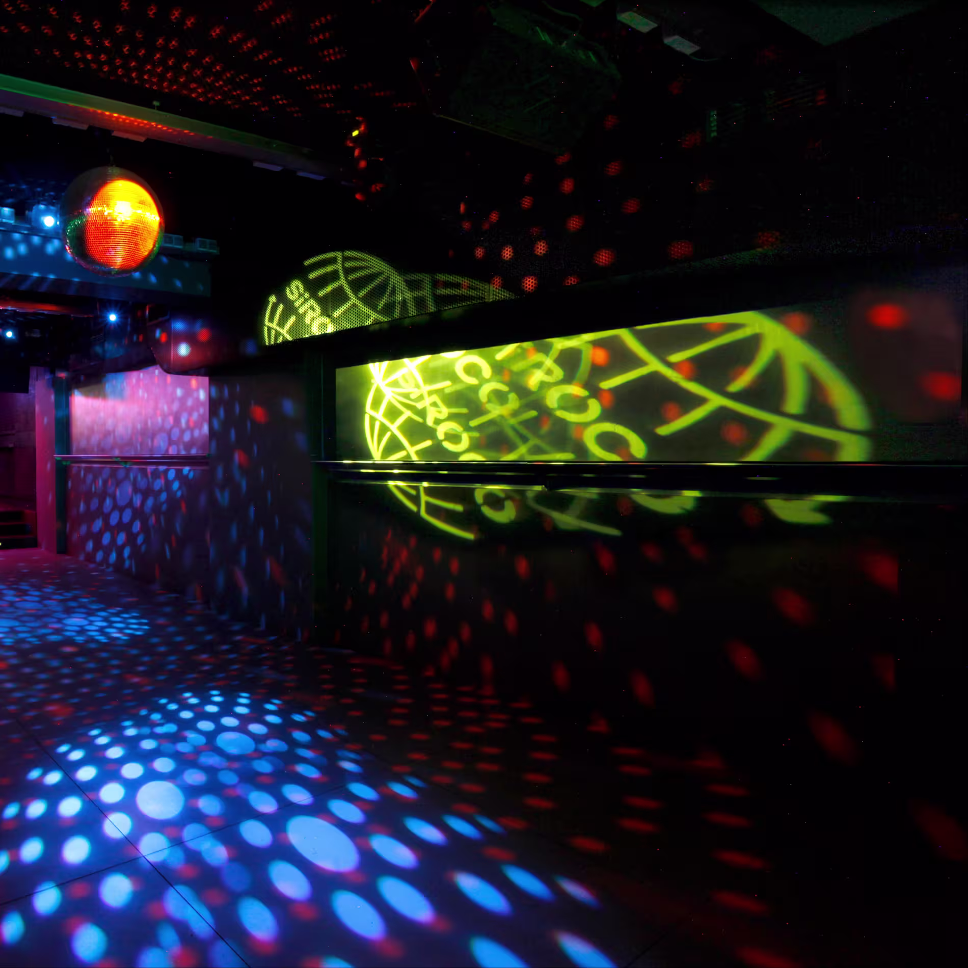 La imagen muestra una pista de baile de la sala Sirocco vacía con varias luces y patrones coloridos proyectados sobre ella. El entorno es una discoteca o un lugar de fiesta, con una bola de discoteca colgando del techo y una cabina de DJ en el fondo