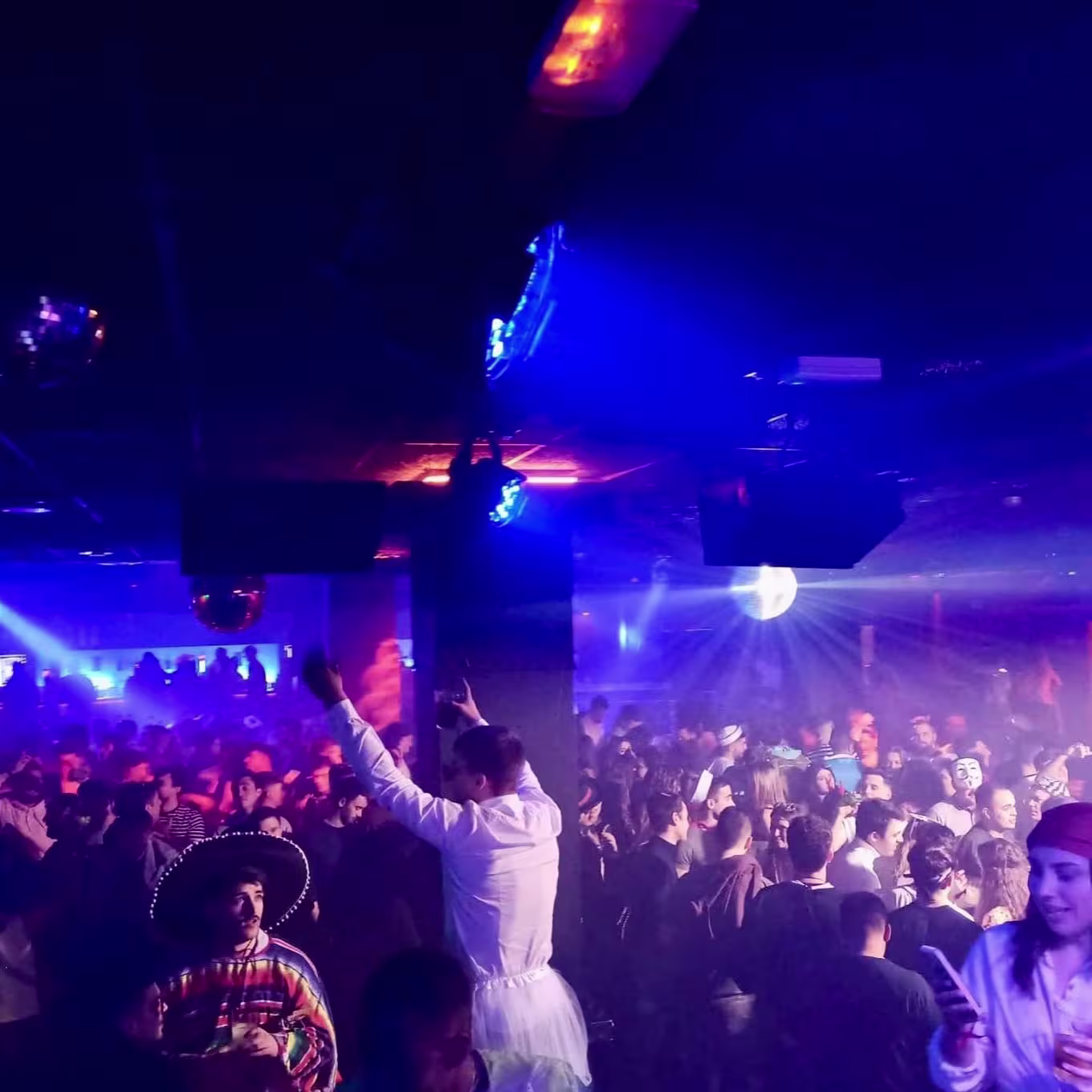 Sala llena en Rock Star con gente disfrazada, luces azules y moradas, bola de espejos girando y ambiente festivo de noche temática.