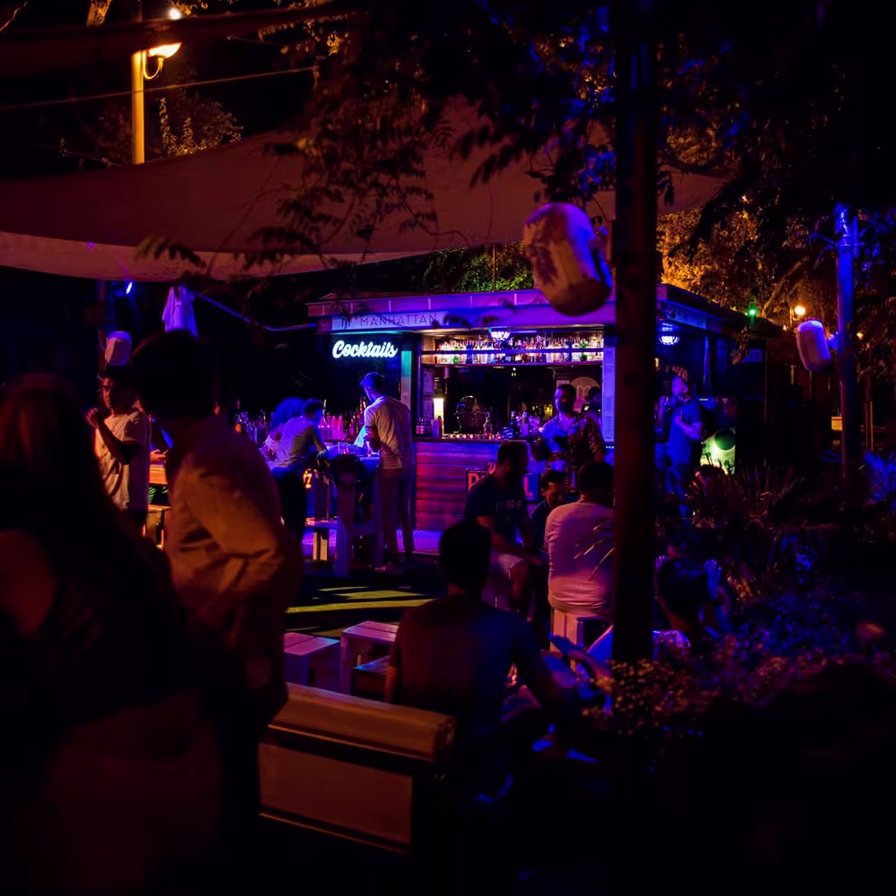 Terraza nocturna de Manhattan River Bar con barra iluminada, cartel de neón “Cocktails” y grupos de personas disfrutando en mesas al aire libre.