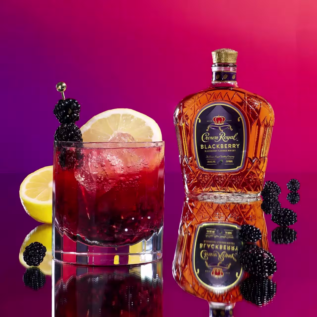 A glass of blackberry cocktail with ice, lemon slice, and fresh blackberries sits next to a bottle of Crown Royal Blackberry whisky on a reflective surface with a purple-red gradient background.