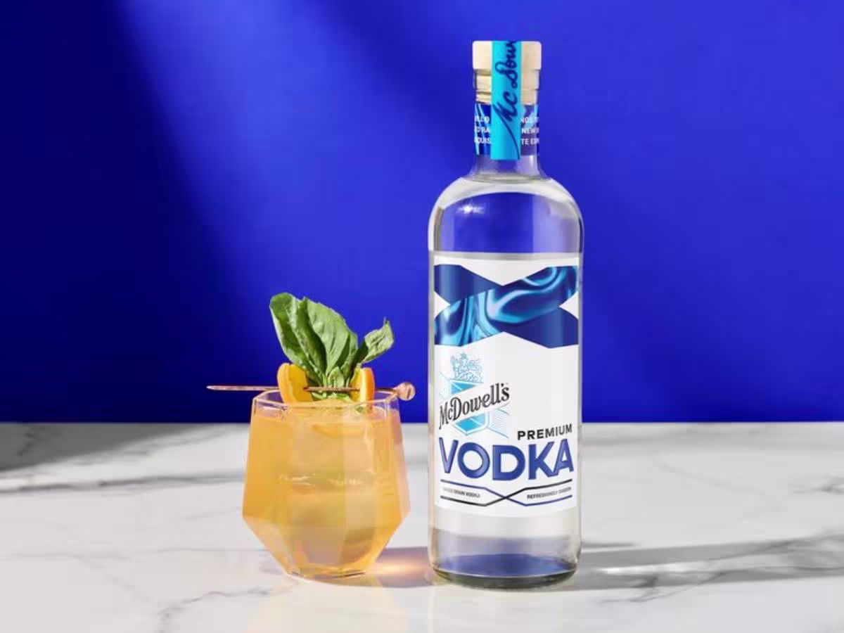 A bottle of McDowells Premium Vodka stands next to a cocktail garnished with herbs and fruit, set on a white marble surface against a blue background.