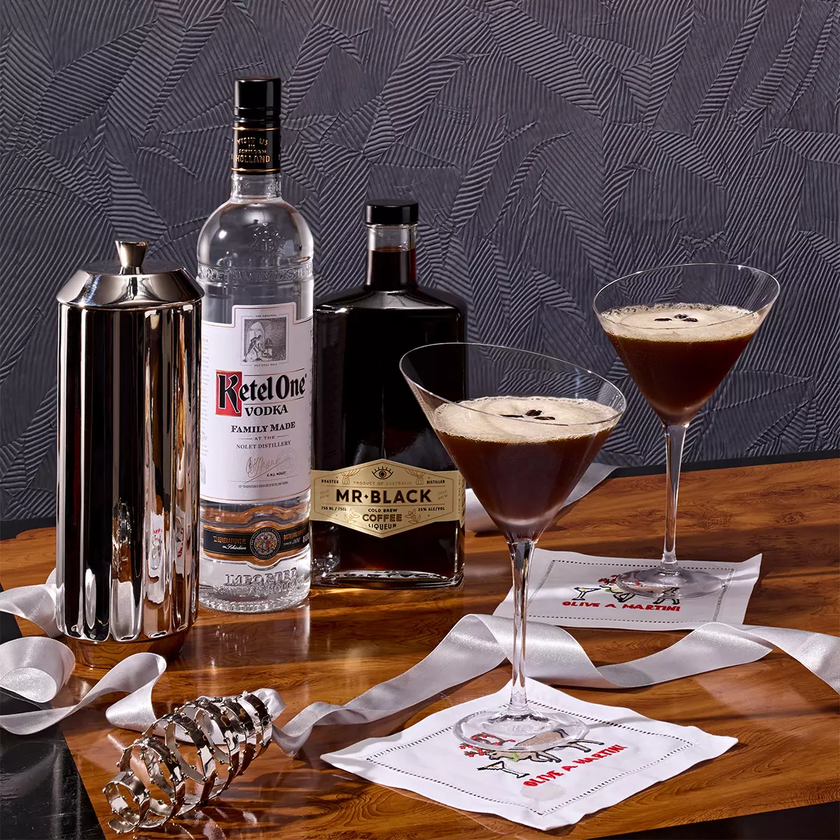 Two espresso martinis in cocktail glasses sit on napkins beside bottles of Ketel One vodka and Mr Black coffee liqueur, a cocktail shaker, and ribbon on a wooden surface.