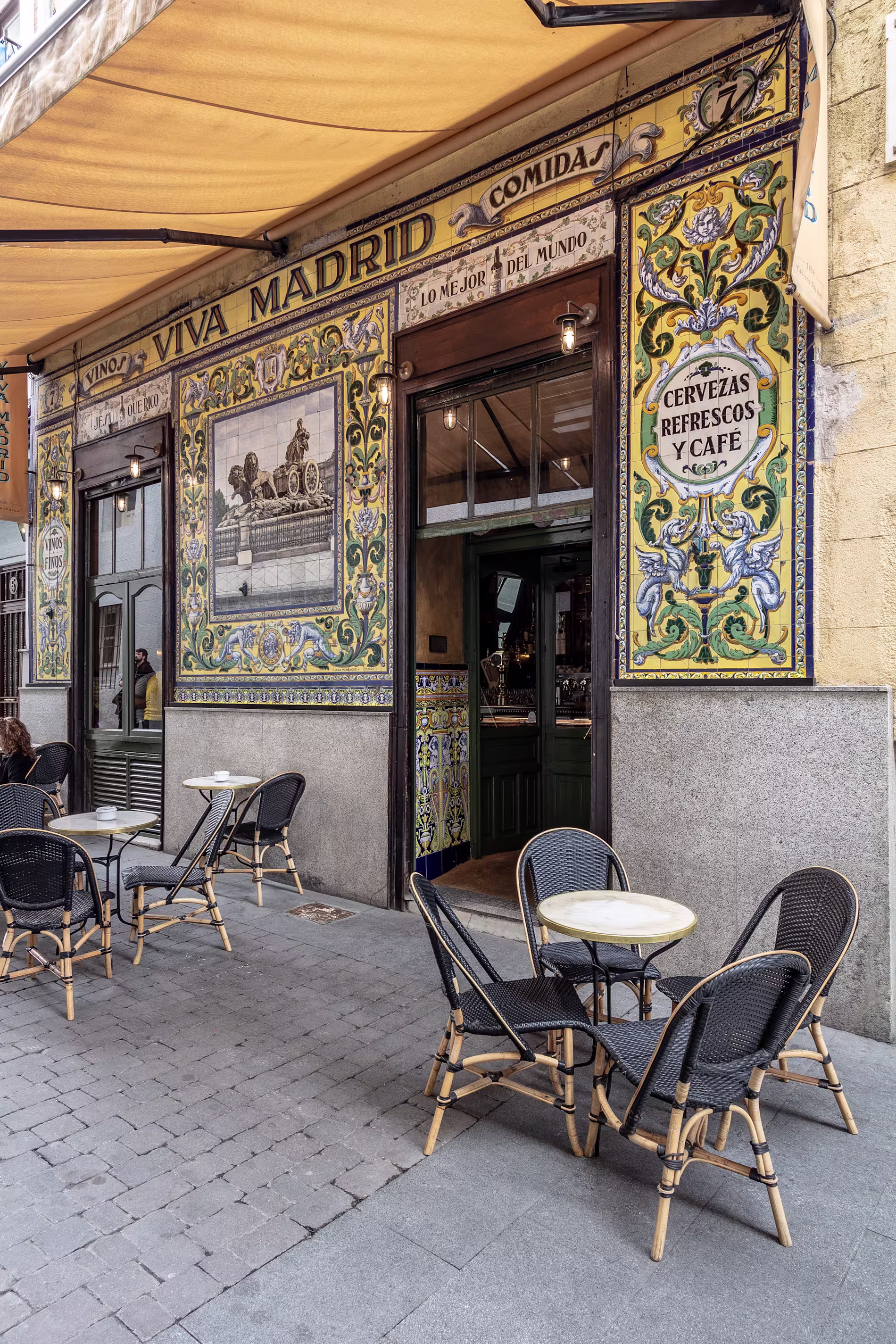 La imagen muestra el exterior del restaurante Viva Madrid  decorada con azulejos coloridos y ornamentados. Los azulejos representan varias escenas y patrones. 
Sobre la puerta, un cartel dice “VIVA MADRID COMIDA”, “CERVEZAS REFRESCOS