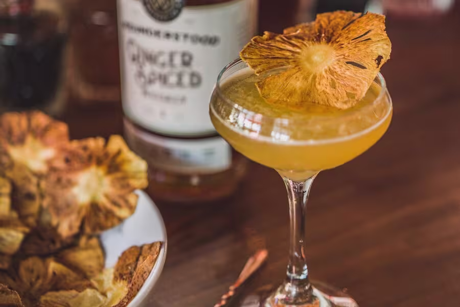 A cocktail in a coupe glass garnished with a dried pineapple slice sits on a wooden table next to a bowl of dried pineapple and a bottle of ginger spiced liquor.