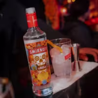 La imagen muestra una botella de vodka Smirnoff Spicy Tamarindo, un vaso con hielo a su lado y un medidor de coctelería.