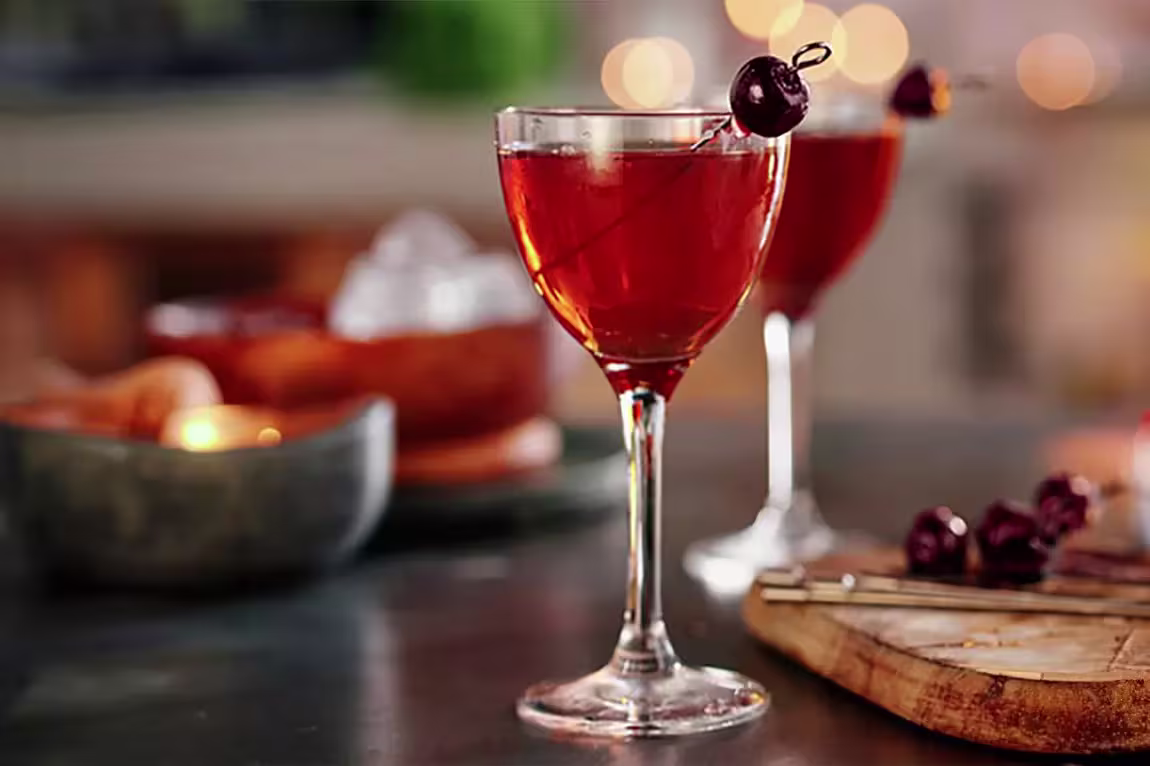 A stemmed glass filled with a red cocktail garnished with two cherries on a pick, with another similar glass and various bar items blurred in the background.