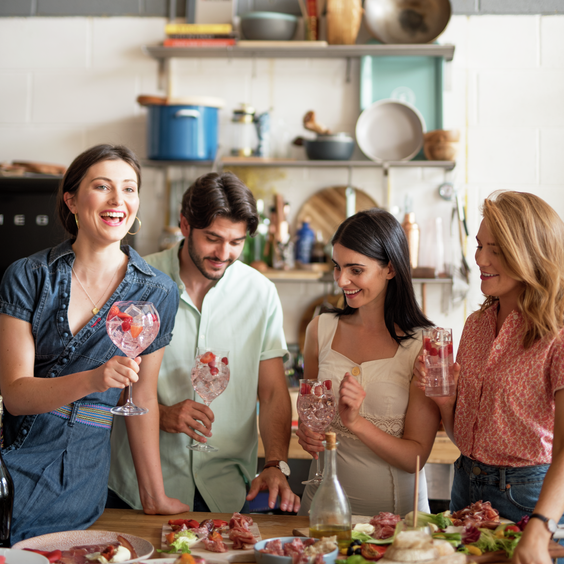 La imagen muestra cuatro personas con distintos cócteles en la mano y una mesa repleta de comida.