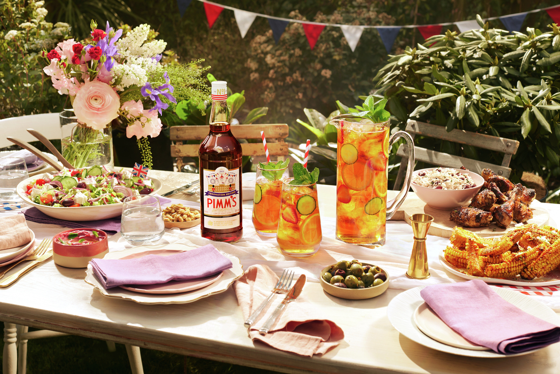 Pimm’s bottle with a pitcher and glasses of Pimm’s cocktail garnished with cucumber and mint on a garden party table with food and flowers