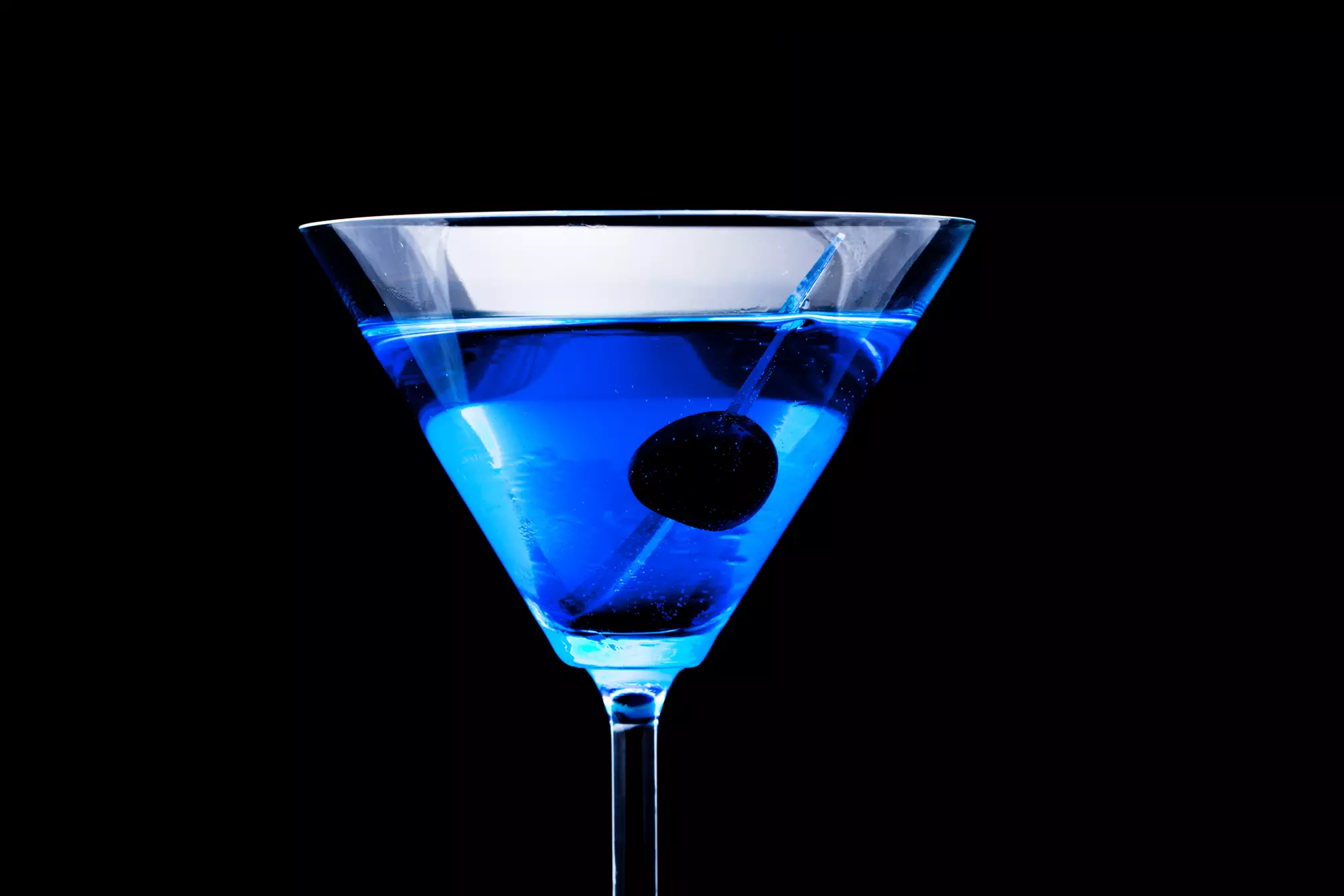 A martini glass filled with bright blue liquid and garnished with a single black cherry, set against a black background.